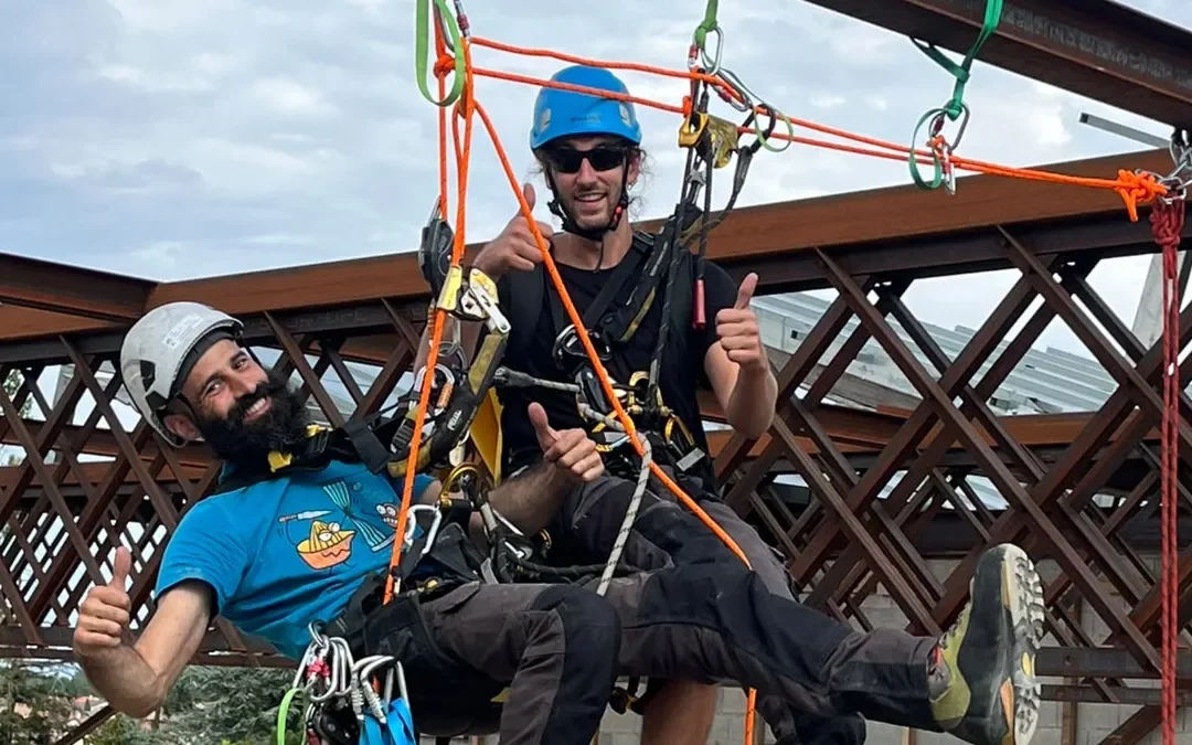 Deux cordistes en formation, pouce levé, suspendus sous une charpente métallique en plein ciel